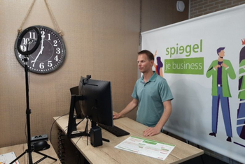 Man staand bij bureau met computer, klok aan de wand, achtergrond met logo van Spiegel je business